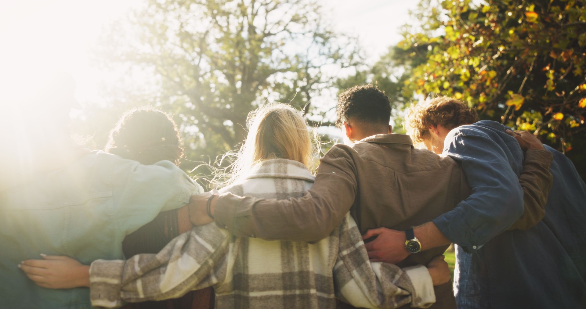 Group, back and friends to relax outdoor on school break, lens flare and hug for bonding in summer. People, students and embrace with support, care and trust together for social reunion at park